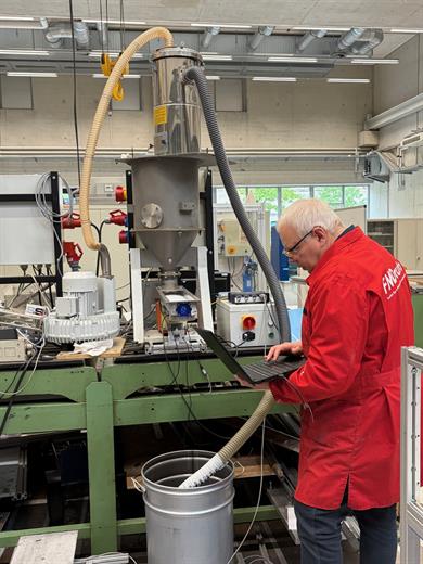 Ein Mann in roter Arbeitsjacke bedient in einem Labor- oder Technikraum einen Laptop vor einer großen Versuchsanlage mit Metallbehälter, Schläuchen und Absaugvorrichtung.